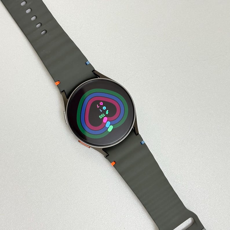 May include: A dark grey smartwatch with a circular face and a matching strap. The watch face shows colourful concentric rings with data. The strap has orange and blue stitching details. A modern wearable tech device.