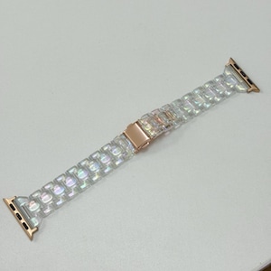 May include: A clear, iridescent Apple Watch band with a rose gold clasp and connectors. The band is made of rectangular, textured links that catch the light. The band is approximately 8 inches long.