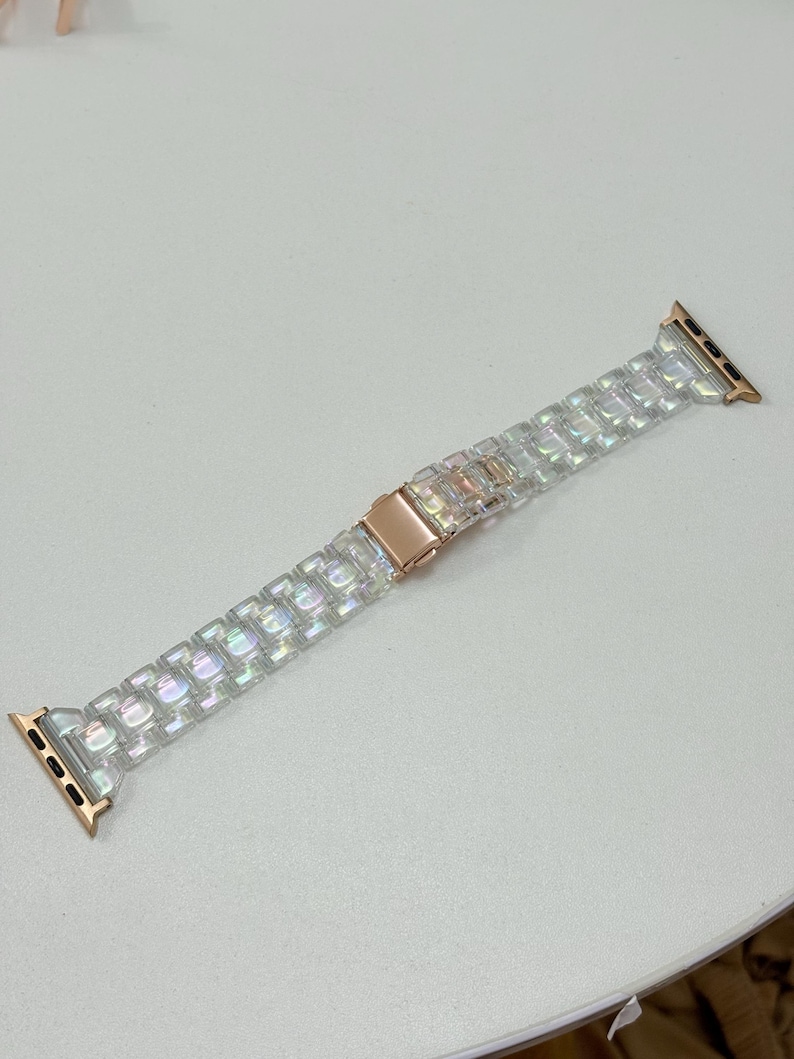 May include: A clear, iridescent Apple Watch band with a rose gold clasp and connectors. The band is made of rectangular, textured links that catch the light. The band is approximately 8 inches long.