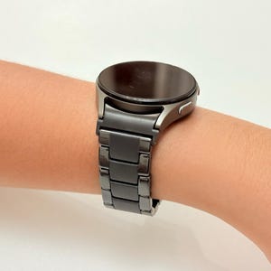 K&ouml;nnte beinhalten: Eine dunkelgraue Smartwatch mit rundem Zifferblatt und passendem Metallarmband. Die Uhr wird am Handgelenk getragen und zeigt ihr schlankes, modernes Design. Das Armband besteht aus rechteckigen Gliedern.