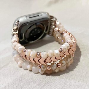 May include: A silver Apple Watch with a rose gold and beaded strap. The strap features multiple strands of off-white and peach-coloured beads, with rose gold chevron-shaped accents. The watch strap is designed to be stylish and decorative.
