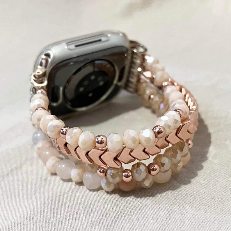 May include: A silver Apple Watch with a rose gold and beaded strap. The strap features multiple strands of off-white and peach-coloured beads, with rose gold chevron-shaped accents. The watch strap is designed to be stylish and decorative.