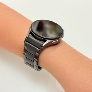 K&ouml;nnte beinhalten: Eine dunkelgraue Smartwatch mit rundem Zifferblatt und passendem Metallarmband wird am Handgelenk einer Person gezeigt. Die Uhr hat ein schlankes, modernes Design und scheint aus langlebigen Materialien gefertigt zu sein. Das Armband besteht aus rechteckigen Gliedern.