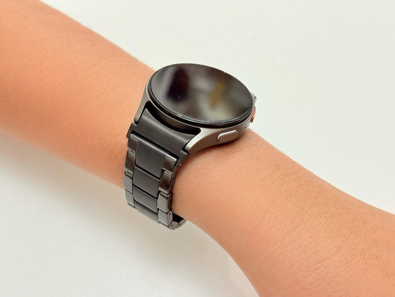 K&ouml;nnte beinhalten: Eine dunkelgraue Smartwatch mit rundem Zifferblatt und passendem Metallarmband wird am Handgelenk einer Person gezeigt. Die Uhr hat ein schlankes, modernes Design und scheint aus langlebigen Materialien gefertigt zu sein. Das Armband besteht aus rechteckigen Gliedern.