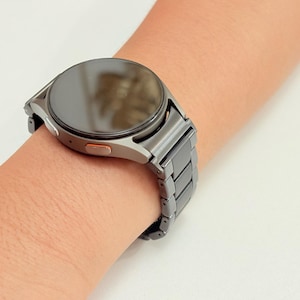 K&ouml;nnte beinhalten: Eine silberne Smartwatch mit dunklem Bildschirm und passendem Metallarmband wird an einem Handgelenk gezeigt. Die Uhr hat ein rundes Zifferblatt mit einem kleinen orangefarbenen Akzent an der Seite. Das Armband besteht aus rechteckigen Gliedern.