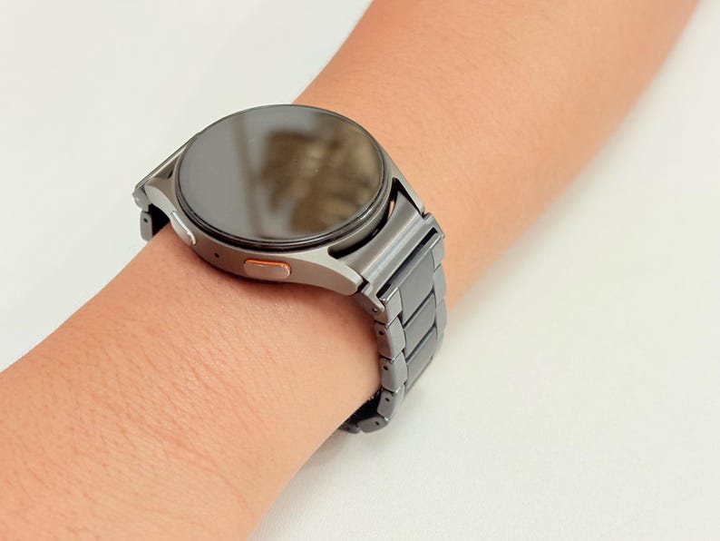 K&ouml;nnte beinhalten: Eine silberne Smartwatch mit dunklem Bildschirm und passendem Metallarmband wird an einem Handgelenk gezeigt. Die Uhr hat ein rundes Zifferblatt mit einem kleinen orangefarbenen Akzent an der Seite. Das Armband besteht aus rechteckigen Gliedern.