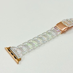 May include: A clear, iridescent Apple Watch band with a rose gold clasp and connectors. The band is made of rectangular, faceted links that catch the light. The watch band is designed to fit an Apple Watch.