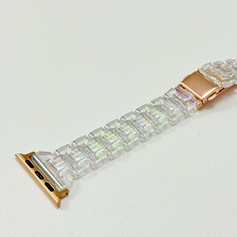 May include: A clear, iridescent Apple Watch band with a rose gold clasp and connectors. The band is made of rectangular, faceted links that catch the light. The watch band is designed to fit an Apple Watch.