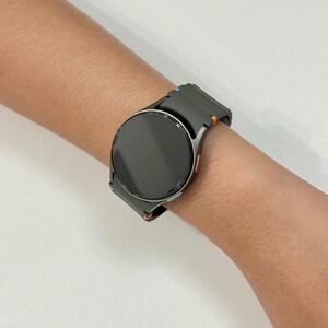 May include: A dark grey smartwatch with a circular face and a matching strap. The strap features orange and blue detailing. The watch is worn on a wrist.