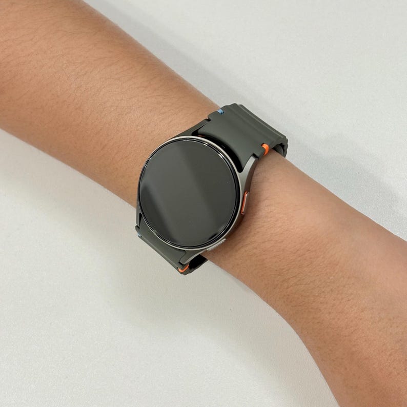 May include: A dark grey smartwatch with a circular face and a matching strap. The strap features orange and blue detailing. The watch is worn on a wrist.