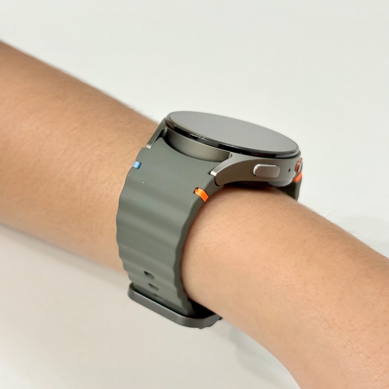 May include: A grey smartwatch with a circular face and a grey strap featuring orange and blue details. The watch is fastened on a wrist.