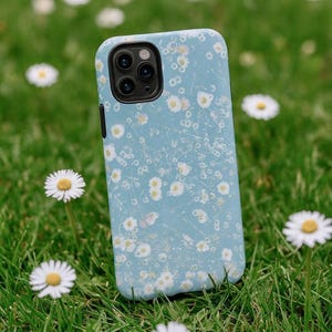 Op de afbeelding: Lichtblauwe telefoonhoes met een wit madeliefjespatroon. De telefoonhoes ligt op groen gras, met verschillende madeliefjes op de achtergrond.