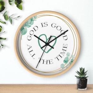 May include: A round wooden wall clock with a white face and gold accents. The clock features a green heart with the text "God is good all the time" in black lettering. The clock is decorated with green leaves.