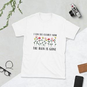 Op de afbeelding: Wit T-shirt met een zwarte afbeelding waarop staat "Ik kan nu duidelijk zien, de regen is weg". De afbeelding bevat ook een rij kleurrijke bloemen.