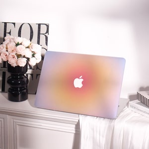 Könnte beinhalten: Ein Laptop mit einer Hülle in Farbverläufen von Rosa, Gelb und Lila. Das Apple-Logo ist weiß. Der Laptop steht auf einer weißen Oberfläche, mit einer schwarzen Vase mit rosa Blumen und einem Buch im Hintergrund.