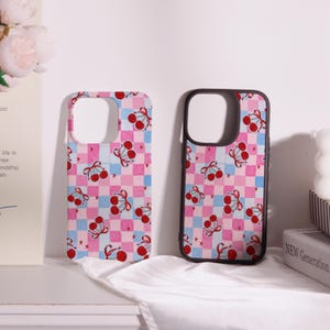 May include: Two phone cases with a pink, blue, and white checkered pattern. Each case features red cherries with bows and small star accents. One case has a black border, while the other has a pink border.