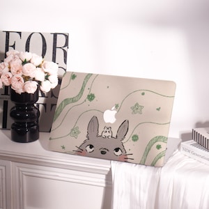 May include: A beige laptop case featuring a cartoon illustration of a gray rabbit with a smaller creature on its head. The case has green wavy lines and star patterns. The Apple logo is visible.