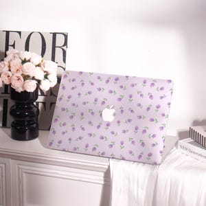 May include: A lavender laptop case with a floral pattern of purple flowers and green leaves. The case has a white Apple logo in the center. The case is on a white surface with a black vase and a book in the background.