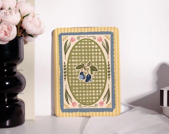 May include: A rectangular decorative card with a yellow and blue striped border. The center features a green and white checkered oval design with a blueberry illustration. Pink floral accents and green leaves complete the design.