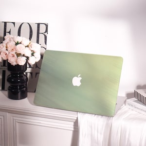Op de afbeelding: Een groene laptophoes met een wit Apple-logo. De hoes heeft een geborsteld metalen uiterlijk. De laptophoes staat op een wit oppervlak naast een zwarte vaas met roze bloemen en een boek.