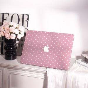 May include: A pink laptop case with a white polka dot pattern. The case features a white Apple logo in the center. The laptop case is on a white surface with a black vase and flowers in the background.