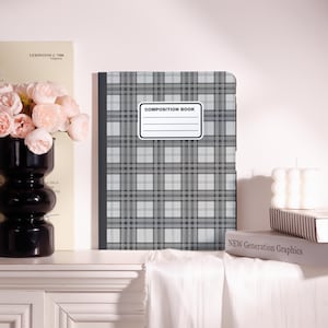 May include: A composition book with a gray and white plaid pattern. The cover features a white label with the words "COMPOSITION BOOK" and lines for writing. The book is displayed with a black vase of pink flowers and a stack of books.