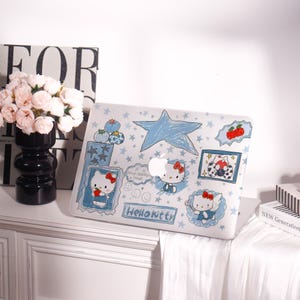 May include: A white laptop case featuring a blue star and Hello Kitty illustrations. The case has a repeating star pattern and several framed drawings of Hello Kitty with bows, blueberries, and strawberries. The words "Hello Kitty" are also visible.