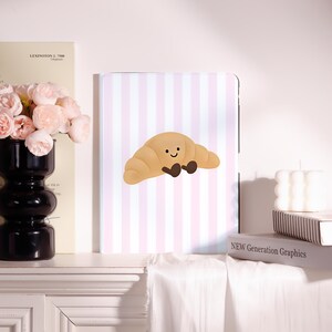 May include: A rectangular tablet case featuring a light pink and white vertical striped pattern with a central graphic of a tan croissant character with a smiling face and small dark brown feet, centered on a white background.