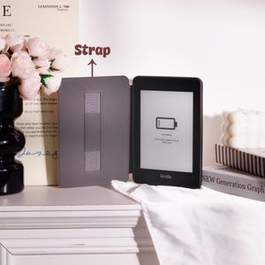 May include: A black Kindle e-reader with a gray cover, open to reveal the screen. The screen displays a low battery icon. The cover has a gray strap. The background includes a vase of pink flowers and a stack of books.