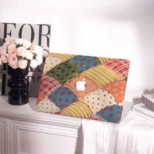 May include: A colorful laptop case with a patchwork design. The case features a variety of patterns, including plaid, polka dots, and floral prints, in shades of red, yellow, blue, and green. The Apple logo is visible in the center.