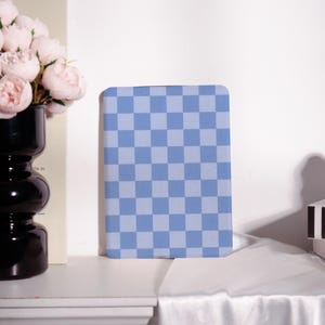Può includere: Un quaderno con un motivo a quadri blu e azzurri. Il quaderno rettangolare ha angoli arrotondati. Lo sfondo include un vaso nero con fiori rosa e una superficie bianca.