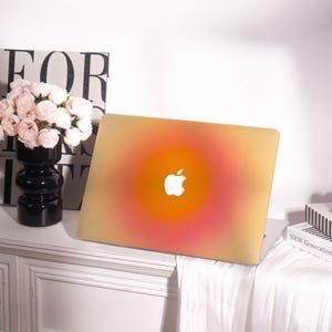 May include: A laptop with a gradient design in shades of yellow, orange, and pink. The Apple logo is visible in white. The laptop is on a white surface, with a black vase of pink flowers and a book in the background.