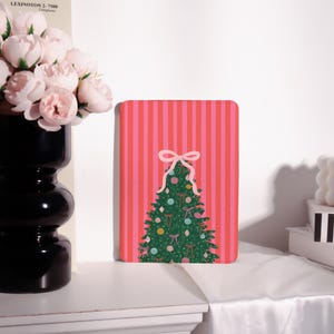 May include: A rectangular Christmas-themed notebook with a pink and red striped background. The notebook features a green Christmas tree decorated with ornaments and bows. A pink bow is at the top of the tree.