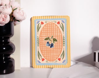 May include: A rectangular decorative card with a yellow and blue striped border. The centre features an oval with a peach gingham pattern and a blueberry illustration. Red and green floral accents surround the oval.