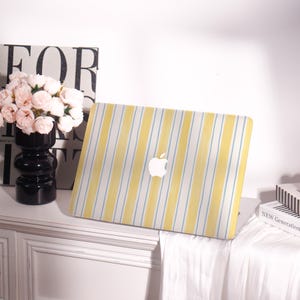 May include: A laptop case with a yellow and white striped pattern and blue accent stripes. The Apple logo is visible in white. The case is on a white surface. A black vase with pink flowers is to the left of the laptop.