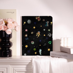 May include: A black tablet case featuring a space-themed pattern with yellow stars, various colorful buttons, and small cartoon character illustrations. The design includes a central figure in a yellow coat, a small mouse, and other whimsical figures.