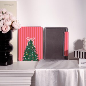 May include: A Christmas-themed tablet case with a pink and red striped background. The case features a green Christmas tree design with ornaments and a white bow. The case is open, revealing a black interior.