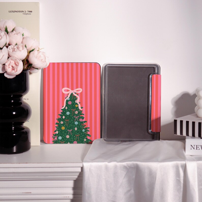 May include: A Christmas-themed tablet case with a pink and red striped background. The case features a green Christmas tree design with ornaments and a white bow. The case is open, revealing a black interior.