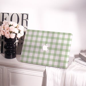 May include: A green and white gingham pattern laptop case with the Apple logo. The case is on a white surface, next to a black vase with pink flowers and a book with the words "NEW GENERATION".
