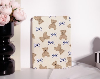 May include: A rectangular cream-colored case featuring a repeating pattern of brown teddy bears and blue bows. The case is upright and appears to be a book cover or a tablet case. The background is a plain white surface.