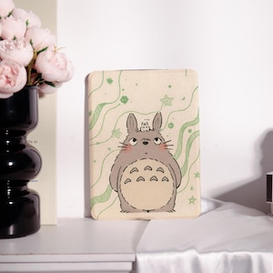 May include: A beige rectangular case featuring a cartoon illustration of Totoro, a large gray forest spirit, with a smaller creature on its head. The background has green wavy lines and stars. The case is likely for a phone or tablet.