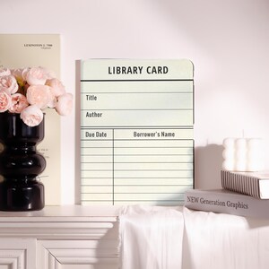 May include: A vintage-style library card with the words "LIBRARY CARD" at the top. The card has spaces for title, author, due date, and borrower's name. The card is cream-colored with black text. The card is displayed on a white surface.