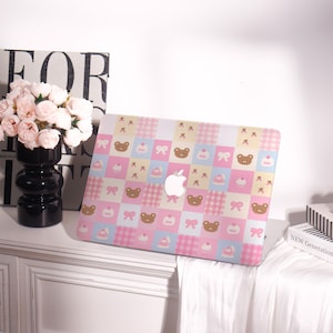 May include: A pink, blue, and yellow patterned laptop case featuring a repeating design of teddy bears, bows, and cupcakes. The case has a white Apple logo cutout. The case is on a white surface.