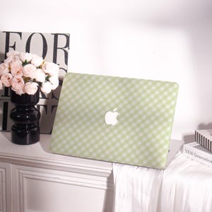 May include: A green and white gingham pattern laptop case with a white Apple logo. The case is on a white surface, next to a black vase with pink flowers and a book with the words "NEW GENERATION".