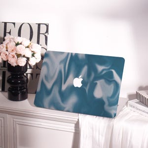 May include: A teal laptop case with a white marble design and a white Apple logo. The case is on a white surface with a black vase of pink flowers and a book with the word "FOR" on it.
