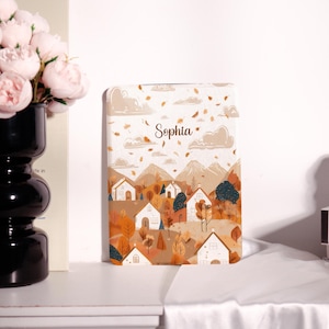 May include: A notebook with a landscape illustration featuring houses, trees, and mountains in shades of brown, orange, and white. The name "Sophia" is written in cursive at the top. The design evokes a fall or autumn theme.