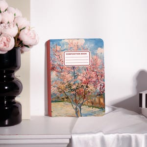May include: A composition book featuring a painting of a blossoming tree with pink flowers against a blue sky. The cover has a red spine and the words "COMPOSITION BOOK" printed on the top. The notebook is on a white surface.