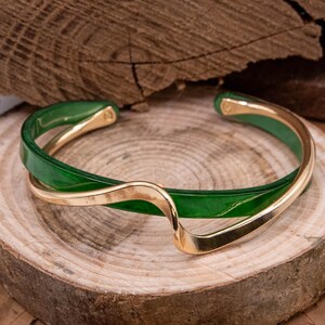 May include: A green and gold-tone bracelet. The bracelet has a glossy green band intertwined with a gold-coloured, wave-shaped accent. The jewellery is displayed on a wooden surface.