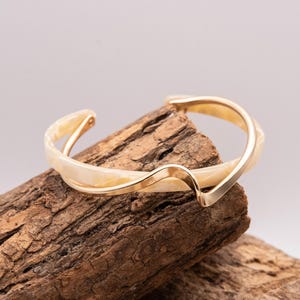 May include: A gold-toned cuff bracelet with a cream-colored, marbled design. The bracelet has a wave-like shape and is displayed on a piece of textured, brown wood. Jewelry.
