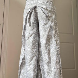 May include: A light gray wrap-style palazzo pants with a white floral pattern. The pants are tied at the waist with a matching fabric tie. A white halter top is visible above the pants.